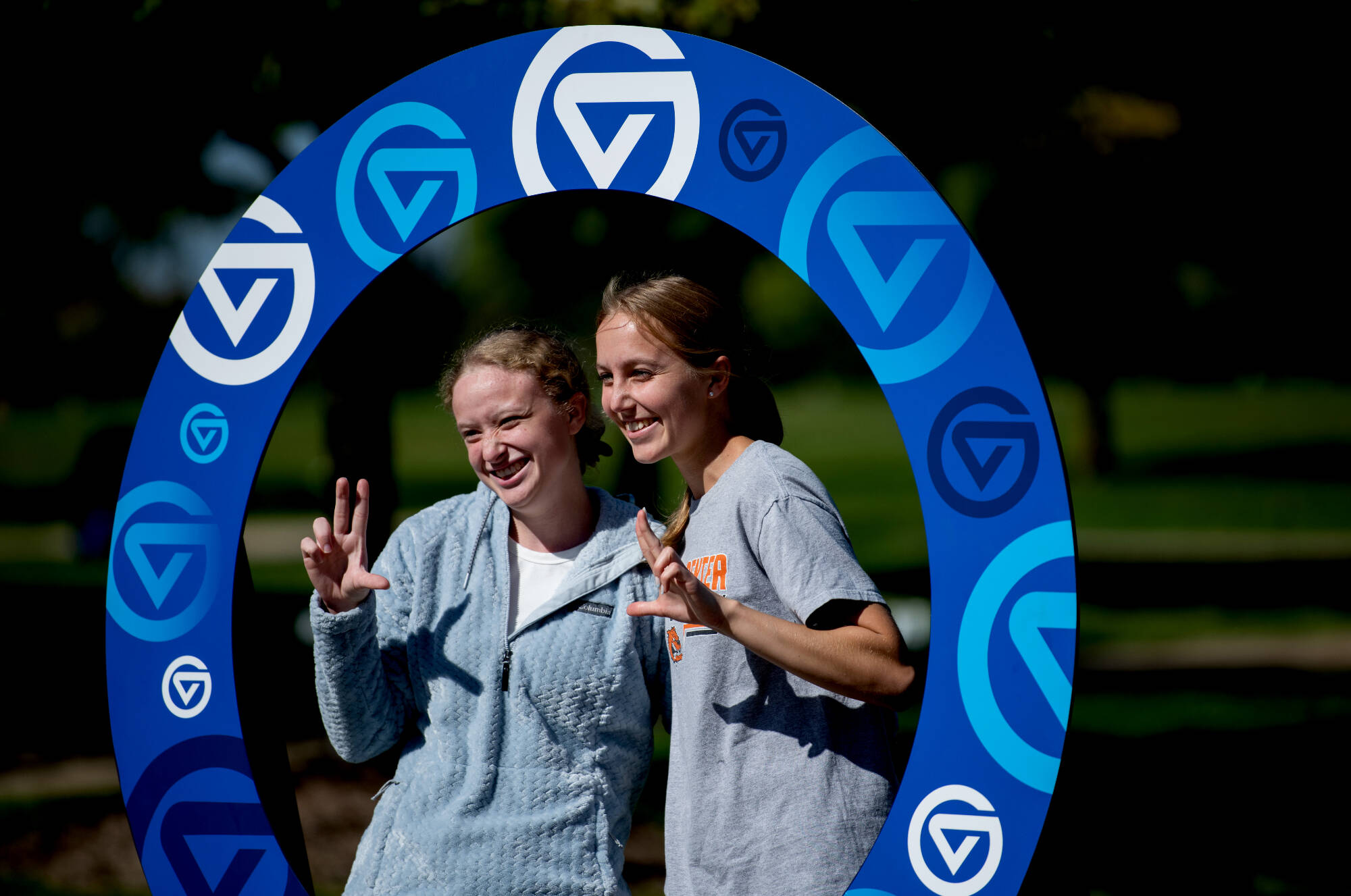 Two smiling students in gray GVSU shirts pose making V-hand gestures through large circular blue photo frame decorated with GVSU logos at outdoor campus event.